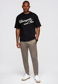 Mężczyzna stoi w czarnej koszulce "Chiringuito and Sun", beżowych spodniach dresowych, białych sneakersach oraz czarnym zegarku na białym tle.