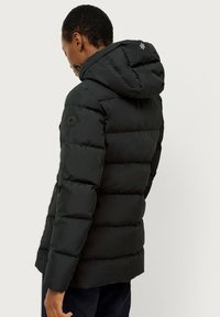 Sort pufferjakke med hætte, prydet med quiltet design, et sort rundt logo på ærmet og lynlåslukning foran.