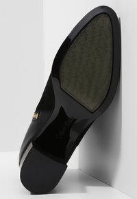 Tacón de cuero negro con parte superior de ante, punta afilada, acento dorado y suela de goma texturizada que presenta un patrón de logo repetido.