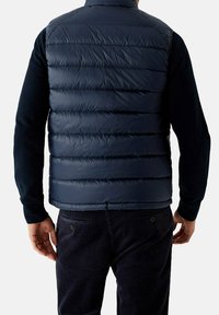 Marineblaue gesteppte Weste mit horizontalen Steppnähten, hohem Kragen und leichtem, glänzendem Finish. Wird über einem marineblauen Langarmshirt getragen.