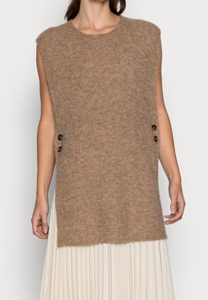 Gilet marron en tricot sans manches avec deux boutons latéraux porté sur une jupe blanche plissée, montré du cou aux hanches.