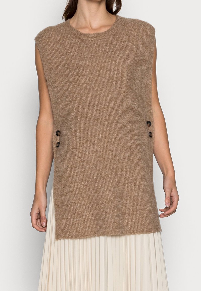 Gilet marron en tricot sans manches avec deux boutons latéraux porté sur une jupe blanche plissée, montré du cou aux hanches.