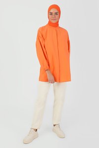 Tunic arancione con colletto alto e maniche arricciate, abbinata a pantaloni dritti beige chiaro e sneaker neutre.