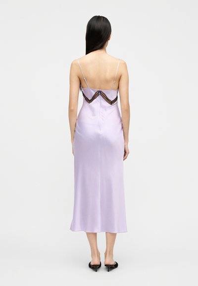 Γυναίκα με μακριά μαλλιά φορεί ένα λιλά σατέν φόρεμα με λεπτομέρειες από μαύρη δαντέλα στην πλάτη και μαύρα παπούτσια με μεσαίο τακούνι, στέκεται μπροστά σε λευκό φόντο.