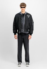 Chaqueta bomber negra con forro acolchado, sudadera con capucha gris, pantalones de mezclilla negros y zapatillas negras con suelas blancas. Lleva puestas gafas de sol cuadradas.