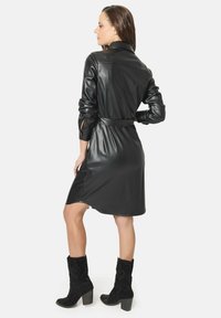 Vestido camiseros de imitación cuero negro con mangas largas, cintura ajustada y aberturas laterales. Largo por la rodilla, textura suave y diseño con cuello.