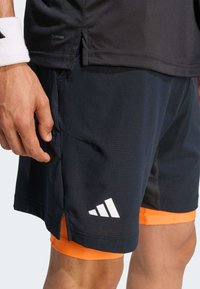 Shorts de sport noirs avec des accents orange, fabriqués en tissu léger. Ils présentent un logo blanc, une fente latérale et une doublure intérieure pour plus de confort.