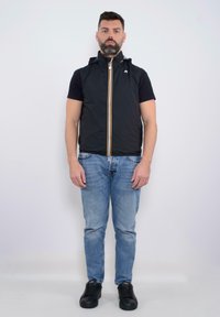 Gilet nero senza maniche con colletto alto, zip frontale con dettagli arancioni, indossato sopra una maglietta nera, abbinato a jeans azzurri chiari e scarpe nere.