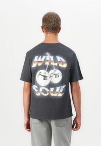 Man with short hair wearing a dark gray T-shirt with chrome cherries and the words "WILD SOUL" in gradient colors on the back.