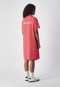 Vestido de camiseta de algodón rosa con mangas cortas, con un logo en la parte posterior y aberturas laterales. Combinado con zapatillas blancas y calcetines de longitud crew.