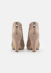 Botines de ante beige con patrones perforados, cremalleras traseras y tacones altos en bloque, mostrados desde la parte trasera sobre un fondo blanco.