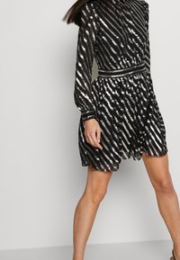 Robe noire courte avec des rayures métalliques argentées, manches longues, taille cintrée et jupe fluide. Le tissu texturé reflète la lumière.