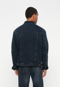 Muškarac u tamnoplavoj traper jakni i odgovarajućim trapericama, viđen sa leđa na jednostavnoj bijeloj pozadini.