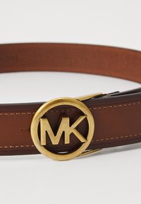 Cintura in pelle marrone con una fibbia circolare dorata che presenta le lettere "MK". Texture liscia e cuciture sottili lungo i bordi.