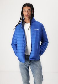 Blauer Puffermantel mit horizontalem Steppmuster, Stehkragen und Logo auf der Brust. Getragen über einem weißen Hemd mit hellblauen Jeans.