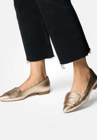 Metalliset kultaiset ballerinat, joissa on terävä kärki, litteä korko ja koristeellinen läppä etuosassa. Sujuva pinta, jossa on kiiltävä viimeistely.