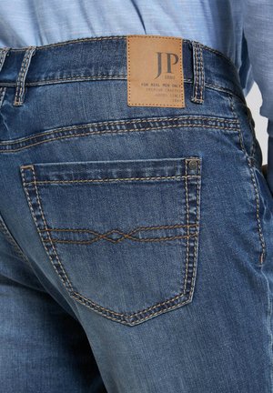 Donkerblauwe denim jeans met een lichtbruin leren label op de tailleband, voorzien van contrasterende bruine stiksels en pocketdetails.
