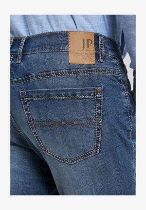 Donkerblauwe denim jeans met een lichtbruin leren label op de tailleband, voorzien van contrasterende bruine stiksels en pocketdetails.