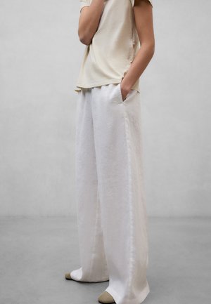 Person iført løse hvide bukser og beige skjorte, stående med den ene hånd i lommen og fødderne i beige slip-on sko på gråt gulv.