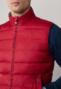 Chaleco acolchado rojo con un cuello alto y líneas de costura verticales. Presenta un cierre frontal y una camisa de manga larga azul marino debajo.