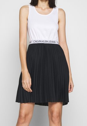 Kobieta w białej bluzce na ramiączkach z logo Calvin Klein Jeans na taśmie oraz czarnej plisowanej spódnicy, stojąca na jednolitym tle.