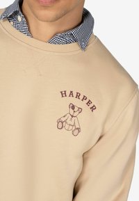 Sudadera beige con un gráfico de un oso y "HARPER" impreso arriba. Camisa con cuello debajo con un patrón de cuadros azul y blanco.