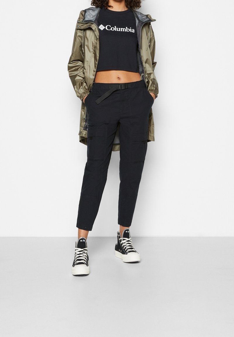 Olivgrön vattentät jacka, svart kortärmad t-shirt med logotyp, svarta cargo-byxor och svarta högklackade sneakers med vita sulor.