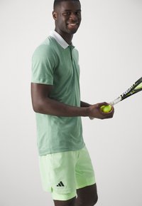 Polo shirt vert chiné avec col blanc, associé à un short vert clair portant un logo Adidas, tenant une raquette de tennis et une balle.