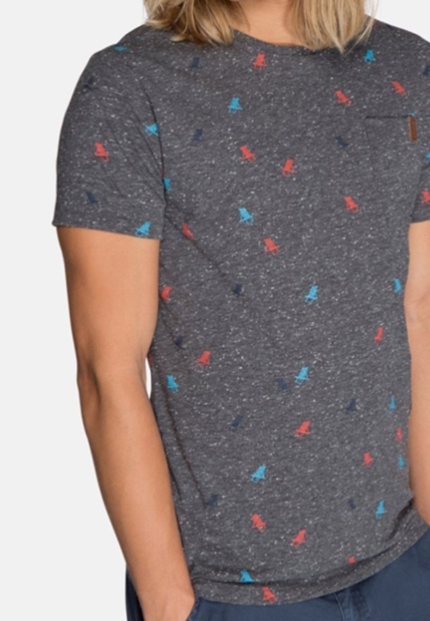 T-shirt à manches courtes gris foncé avec de petits motifs de chaises rouges, bleues et bleu marine, porté par une personne les mains dans les poches.