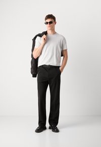 T-shirt blanc à col ras du cou, pantalon noir, ceinture noire et chaussures noires. Modèle tenant une veste noire, portant des lunettes de soleil sombres. Design simple et moderne.