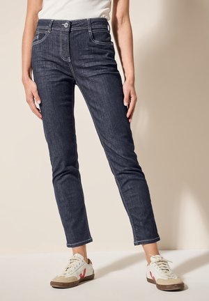 Femme portant un jean slim bleu foncé et des baskets blanches avec des détails beige et rouges, debout sur un fond clair uni.