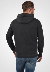 Haut à capuche gris foncé avec une poche kangourou et un capuchon à cordon de serrage, fabriqué en tissu doux. Porté par-dessus un jean bleu avec des coutures visibles.