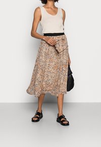 Bloemige midi-rok in beige, oranje en crèmekleuren, gedragen met een licht tanktop. Voorzien van een elastische tailleband en zwarte sandalen.