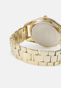 Montre en métal doré avec une surface lisse et polie et un bracelet à maillons. Elle possède un boîtier rond et un fond en acier inoxydable.