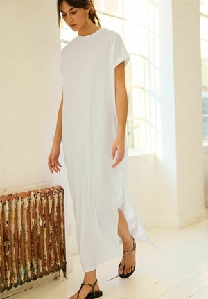 Robe maxi blanche à manches courtes en tissu doux avec une coupe décontractée, présentant des fentes latérales pour plus de mobilité. Associée à des sandales noires plates.