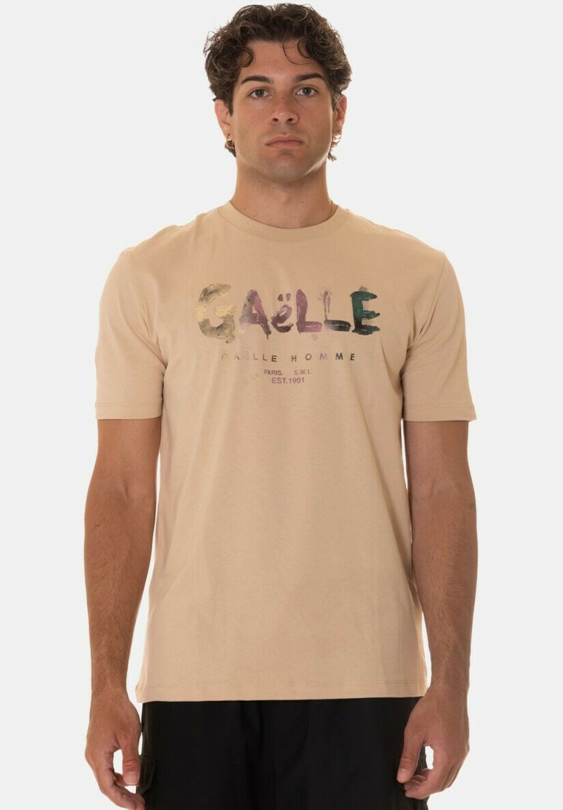 T-shirt in cotone beige con una stampa grafica multicolore che presenta il testo "GAËLLE" e il nome del marchio sotto. Collo girocollo e maniche corte.