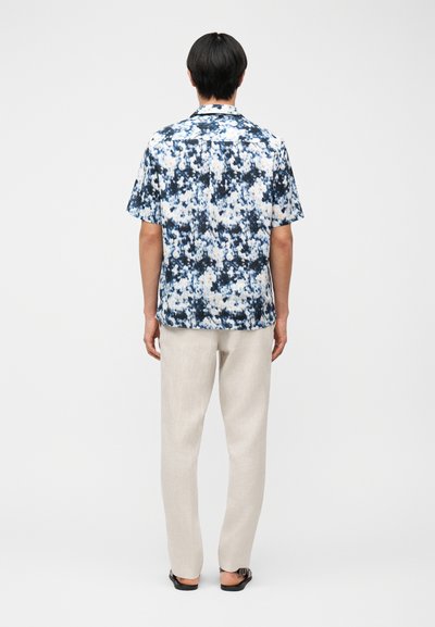 Άνδρας που φοράει κοντομάνικο πουκάμισο με μπλε και λευκό σχέδιο, μπεζ παντελόνι και μαύρα σανδάλια, στέκεται γυρισμένος με την πλάτη προς τα εμπρός σε απλό φόντο.