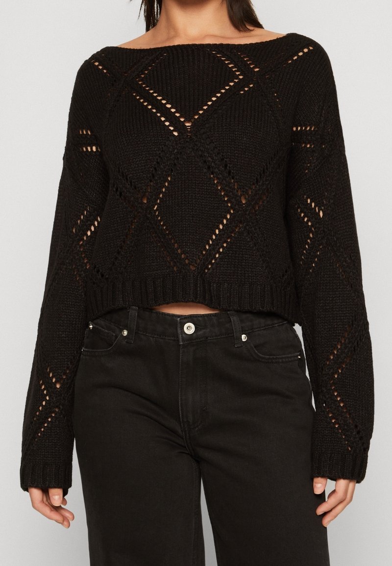 Pull court en maille noire avec un large décolleté, des découpes en forme de losanges et des poignets côtelés, assorti à un jean noir.