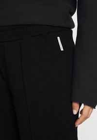 Czarne dresy z elastycznym paskiem w talii, bocznymi kieszeniami i małą białą metką z przodu. Miękki, elastyczny materiał o gładkiej fakturze.
