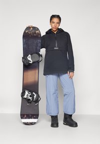 Black snowboard jacket with zipper, paired with light blue snow pants. Holding a dark snowboard with white/black bindings. Black boots.