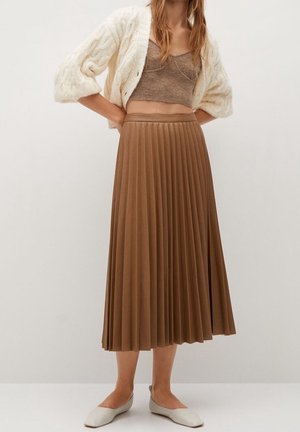 Femme portant un cardigan en maille torsadée crème, un crop top en maille taupe, une jupe midi plissée marron et des chaussures plates blanches pointues, debout devant un fond uni.