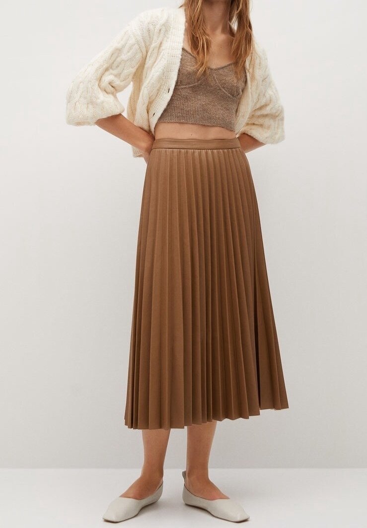 Femme portant un cardigan en maille torsadée crème, un crop top en maille taupe, une jupe midi plissée marron et des chaussures plates blanches pointues, debout devant un fond uni.
