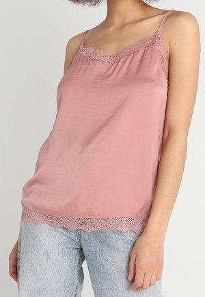 Frau trägt ein staubig rosa Spitzentop mit Spaghettiträgern und hellblaue Jeans, steht vor einem schlichten Hintergrund.