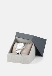 Silver metal watch with white dial, Roman numeral markers, and fluted bezel, displayed in a two-tone gray presentation box.
