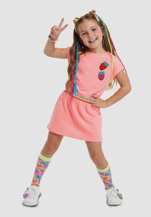 Pige iført lyserød kjole med jordbær-patches, farverigt flettet hår, mønstrede knæstrømper, hvide sneakers, poserer med fredstegn.