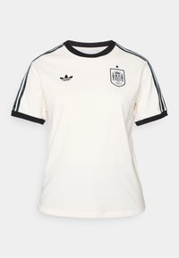 Kremowa koszulka z czarnym kołnierzykiem i mankietami, z czarnym logo Adidasa oraz herbem reprezentacji Hiszpanii, dodatkowo z ciemnymi paskami na rękawach.