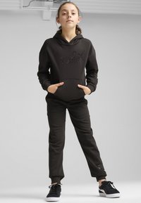 Svart hoodie med präglad logotyp, kängurufickor, matchande svarta joggers och svarta sneakers med vita sulor, mot en neutral bakgrund.