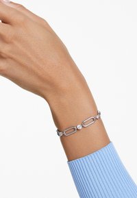 Silverarmband med ovala länkar och runda klara stenar, med en slät yta. Bärs på ett handled med en ribbad blå ärm synlig.