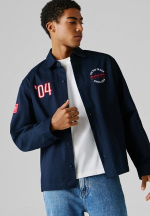 Mladý muž nosiaci námornícku bundu Red Bull Racing cez biele tričko a modré džínsy, pozerajúci sa bokom na obyhlivom sivom pozadí.