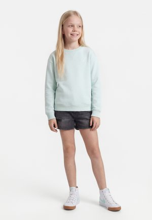 Mladé dievča s dlhými blond vlasmi, oblečené v svetlomodrej mikine, čiernych rifľových kraťasoch a bielych teniskách s vysokým zvrškom, usmieva sa a stojí.